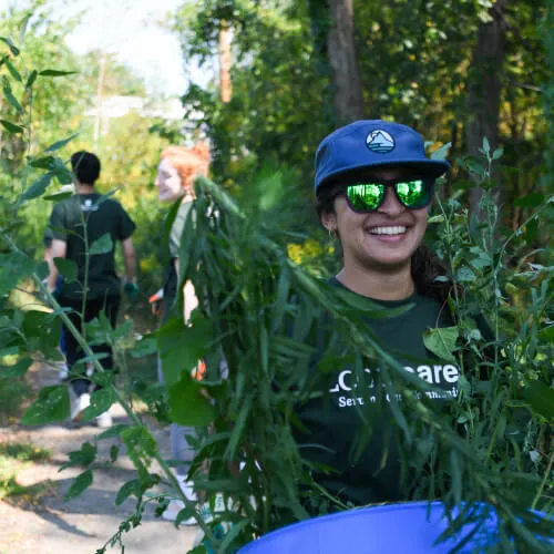 Medical student wearing reflective sunglasses and truckers cap carrying a bin of weeds at a community service location