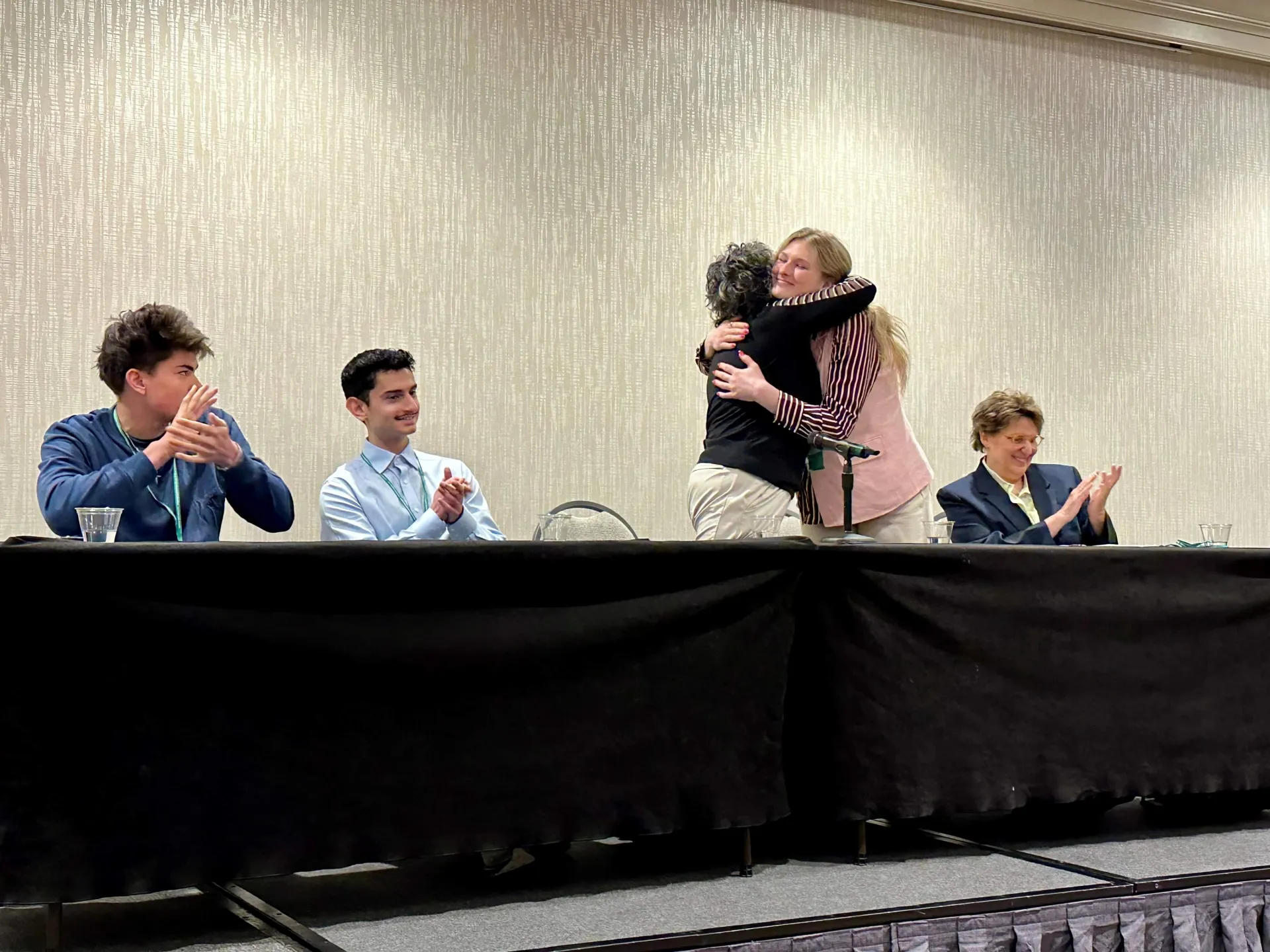 Panelists hugging and clapping at a long table on stage