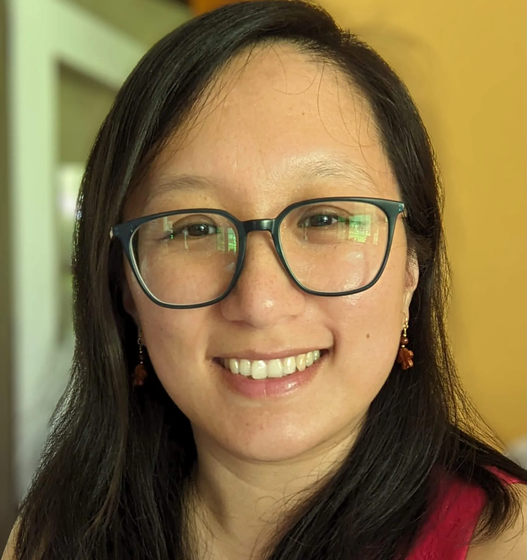 A close-up portrait of a woman wearing glasses, a red top and small red flower earrings, smiling at the camera.