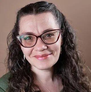 A woman with long, curly dark hair and glasses smiles at the camera. She wears a green top and a silver necklace.