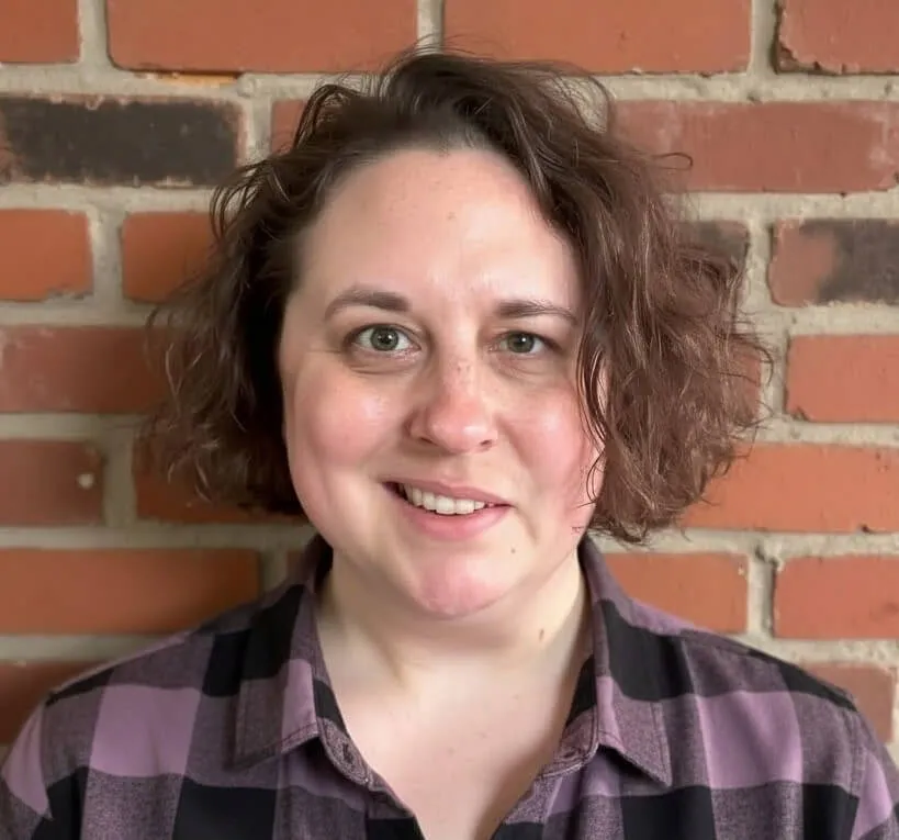 A person with short, wavy hair wearing a purple and black plaid shirt smiles in front of a brick wall.