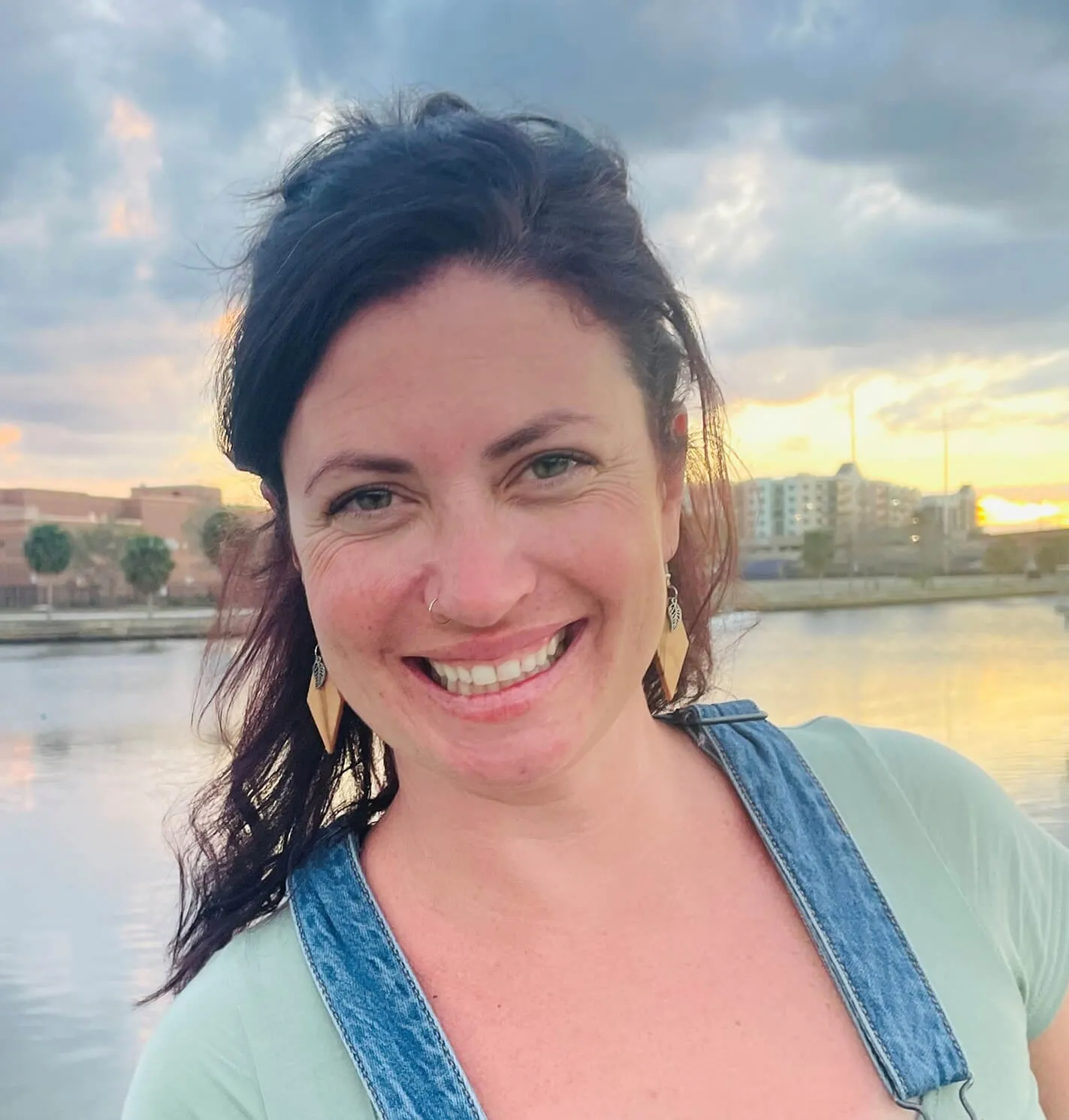 A smiling woman wearing denim overalls and a light green shirt stands in front of a body of water with a city skyline in the background under a cloudy sky at sunset.