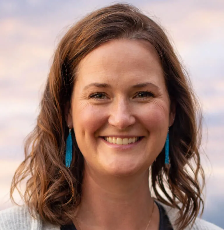 A woman with brown wavy hair and blue tassel earrings smiles warmly. She wears a gray cardigan over a dark top. The sky behind her is a soft pink and orange gradient.