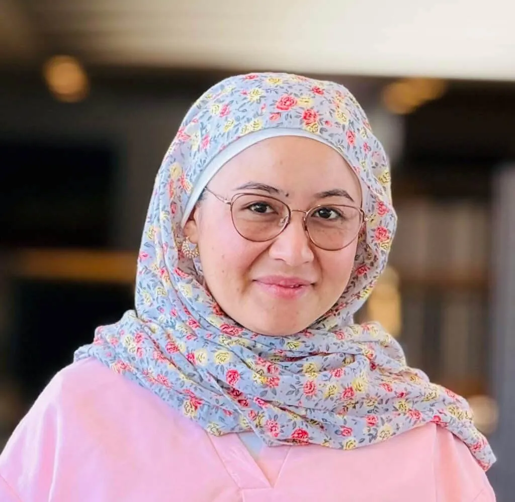 A woman wearing a pink top, a floral hijab, and glasses smiles at the camera.  She wears gold earrings. 