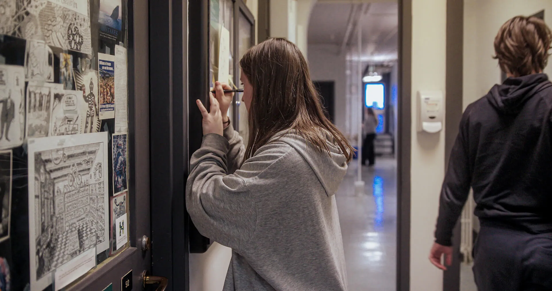 Person writes notes while examining posters and displays on a wall.