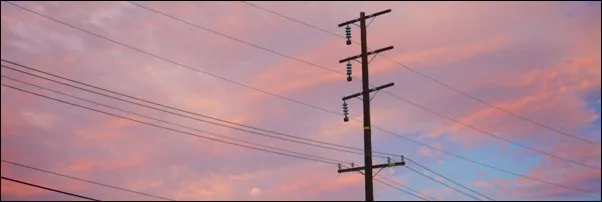 Photograph of a utility pole with multiple power lines against a colorful sunset sky featuring shades of pink, purple, and blue. The scene highlights the contrast between man-made infrastructure and natural beauty during twilight.
