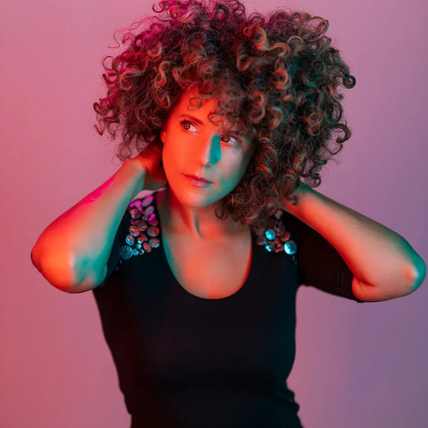 A woman with curly hair poses with her hands behind her neck