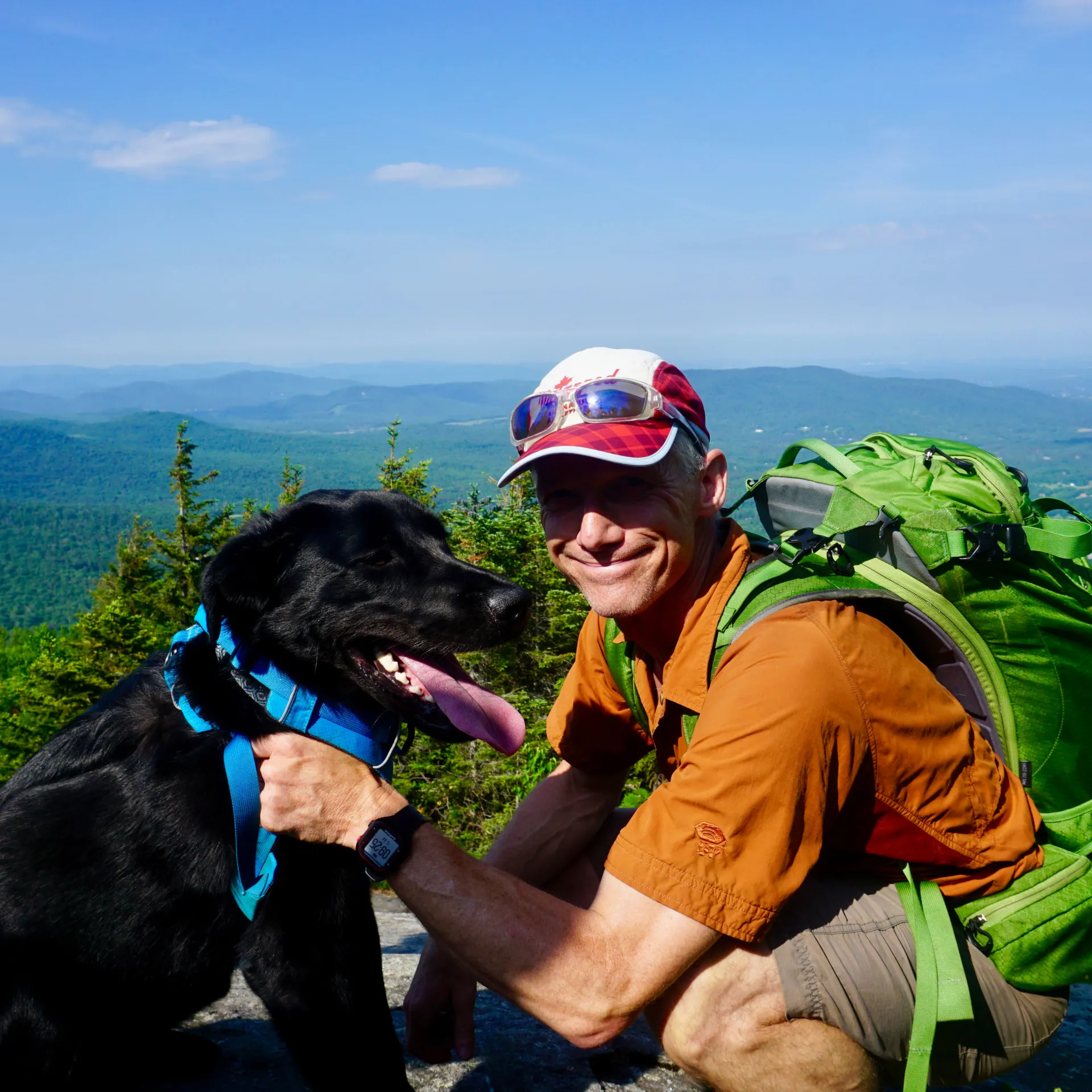 Black dog with tongue out (left) next to crouching, smiling man in hiking gear (right) in front a mountainous landscape.