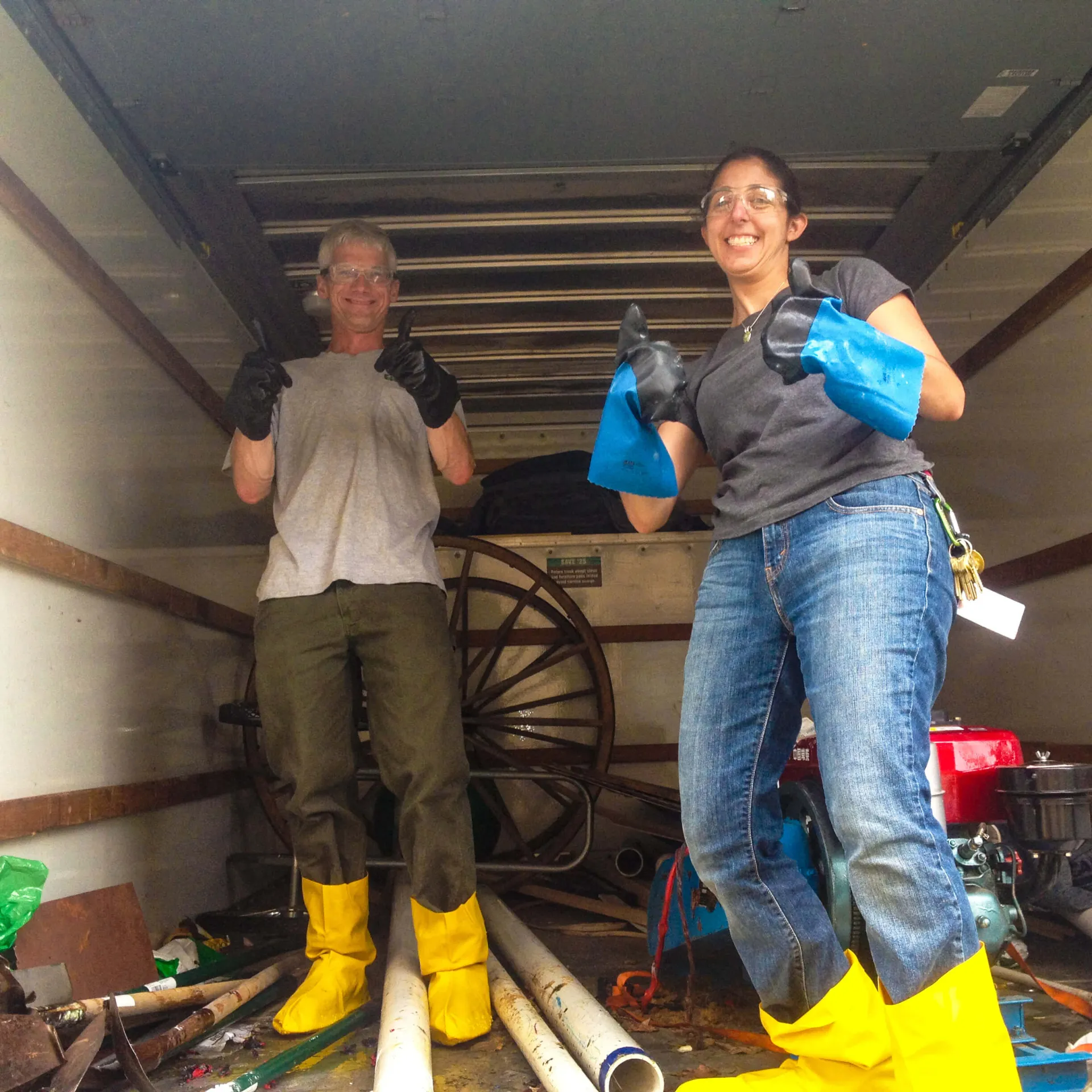 Man (left) and woman (right) wearing safety goggles, rubber gloves, and yellow safety boot covers inside the back of a box truck full of equipment.