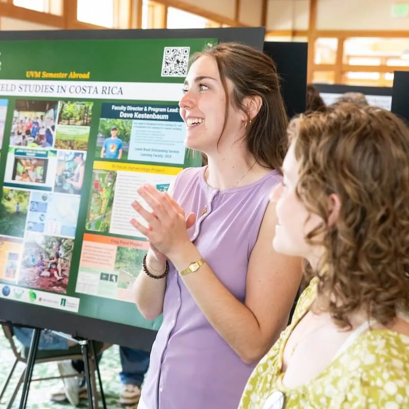 Two students standing next to a research poster, they are smiling and talking to someone out of frame