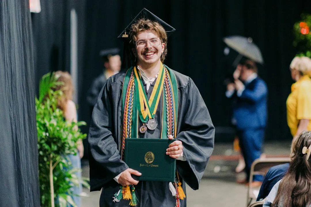 Graduating student Henry Moats wearing a cap and gown and a diploma, they are smiling with multiple cords and metals around their neck.