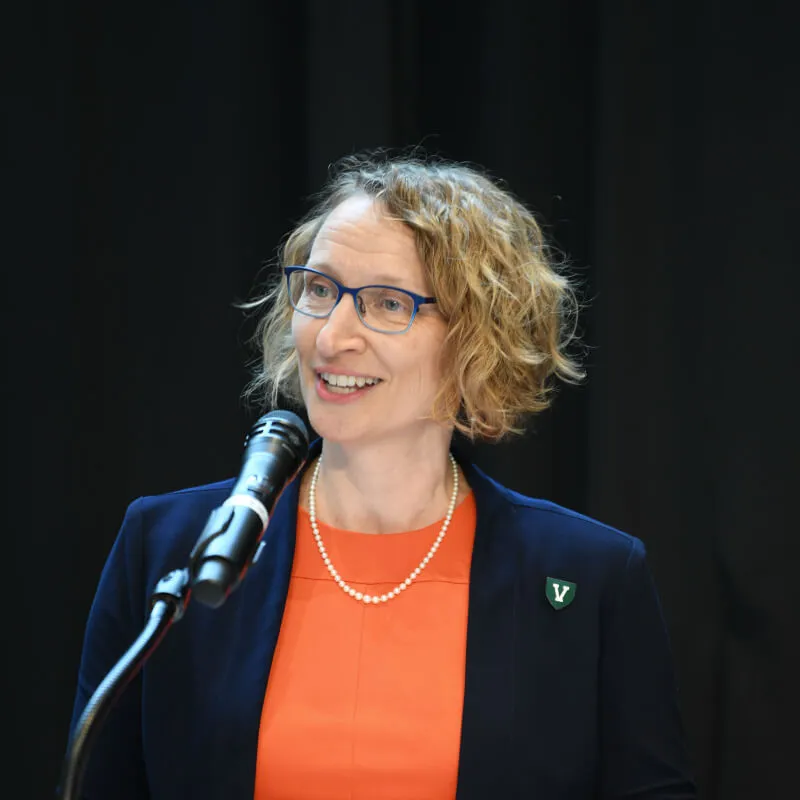 light skinned woman with curly blonde hair, red shirt, dark blazer speaking at a lecturn