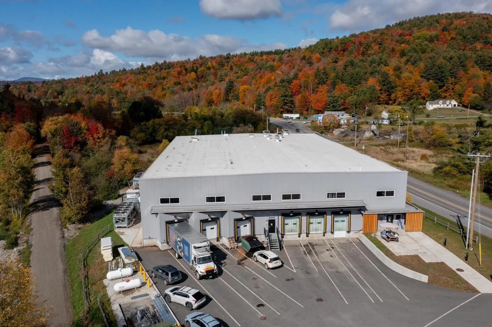 Aerial photograph of a large warehouse with multiple loading docks, several trucks, and cars parked in front. Surrounding area features a road, railway track, and forested hills with autumn foliage under a partly cloudy sky.