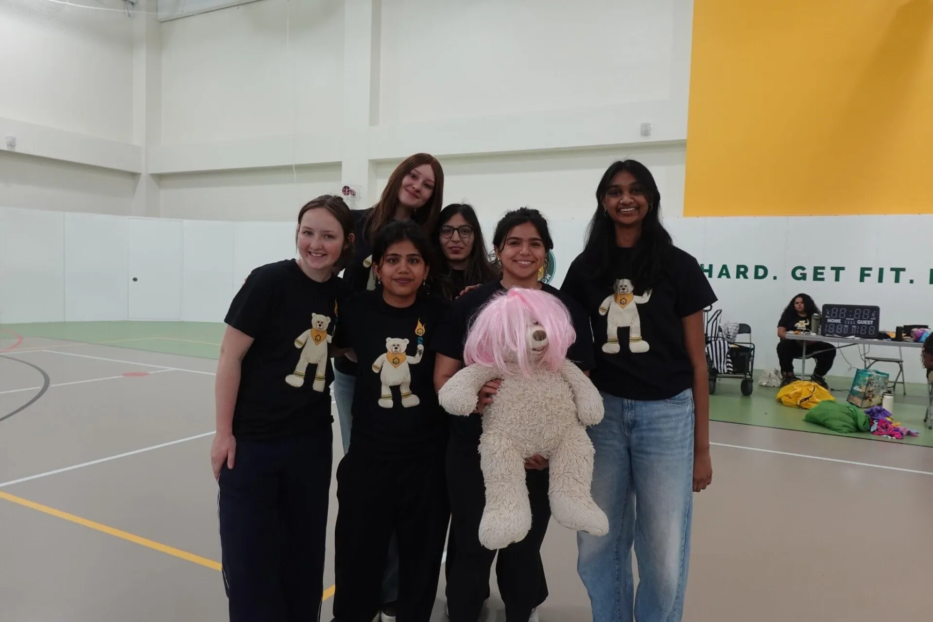 a group of mosaic students in a group huddle all wearing matching Mosaic olympic 2026 shirts with mosaic logo and holding a white oversize teddy bear with a pink wing on it
