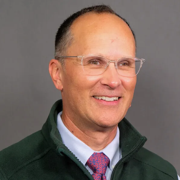 light skinned man with glasses, short gray hair wearing a dress shirt, tie and green fleece vest