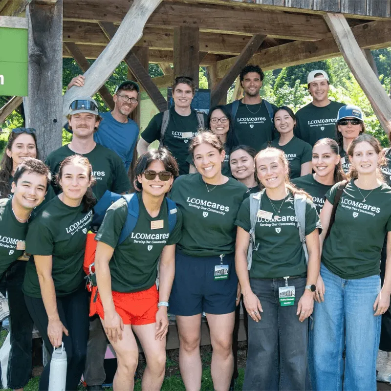 A large group of medical students in green shirts and shorts all in rows smiling at the camera for a group photo. They are outside under a wooden pergola.