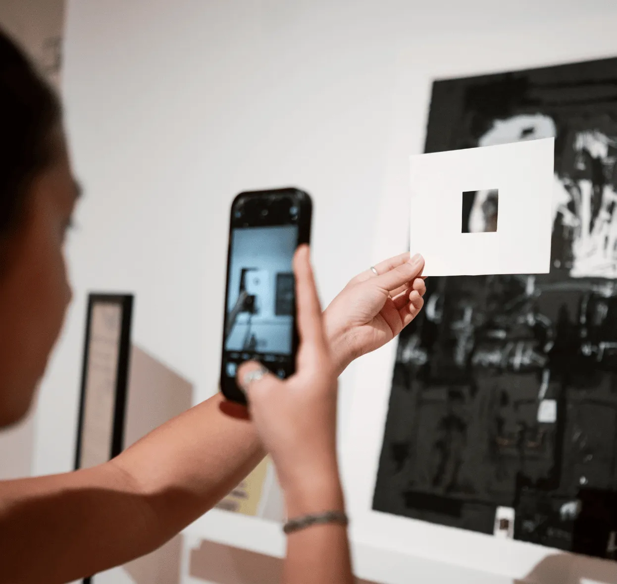 A student takes a photo of a work of art with their phone through a cardboard viewfinder.