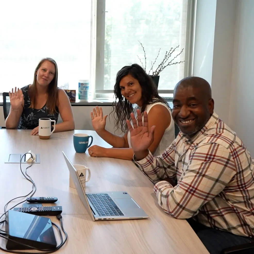two women and a man wave at the camera while seated around a conference table