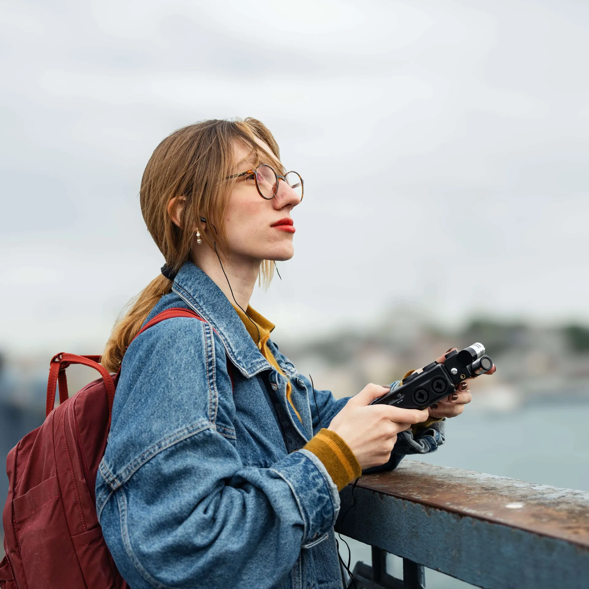 Woman wearing headphones and holding an audio recorder while overlooking a city waterfront.