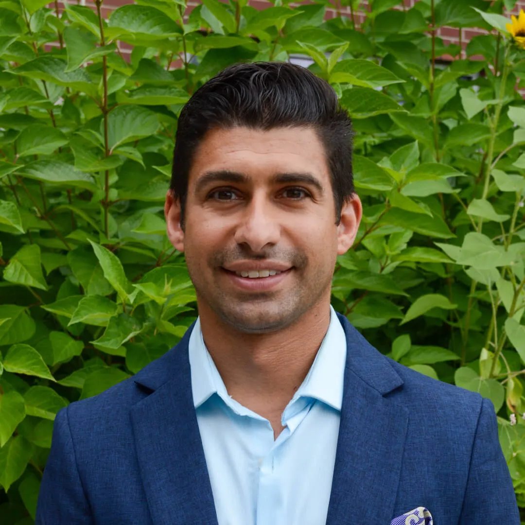 man with short dark hair, blue shirt and navy blazer standing in front of some green plants
