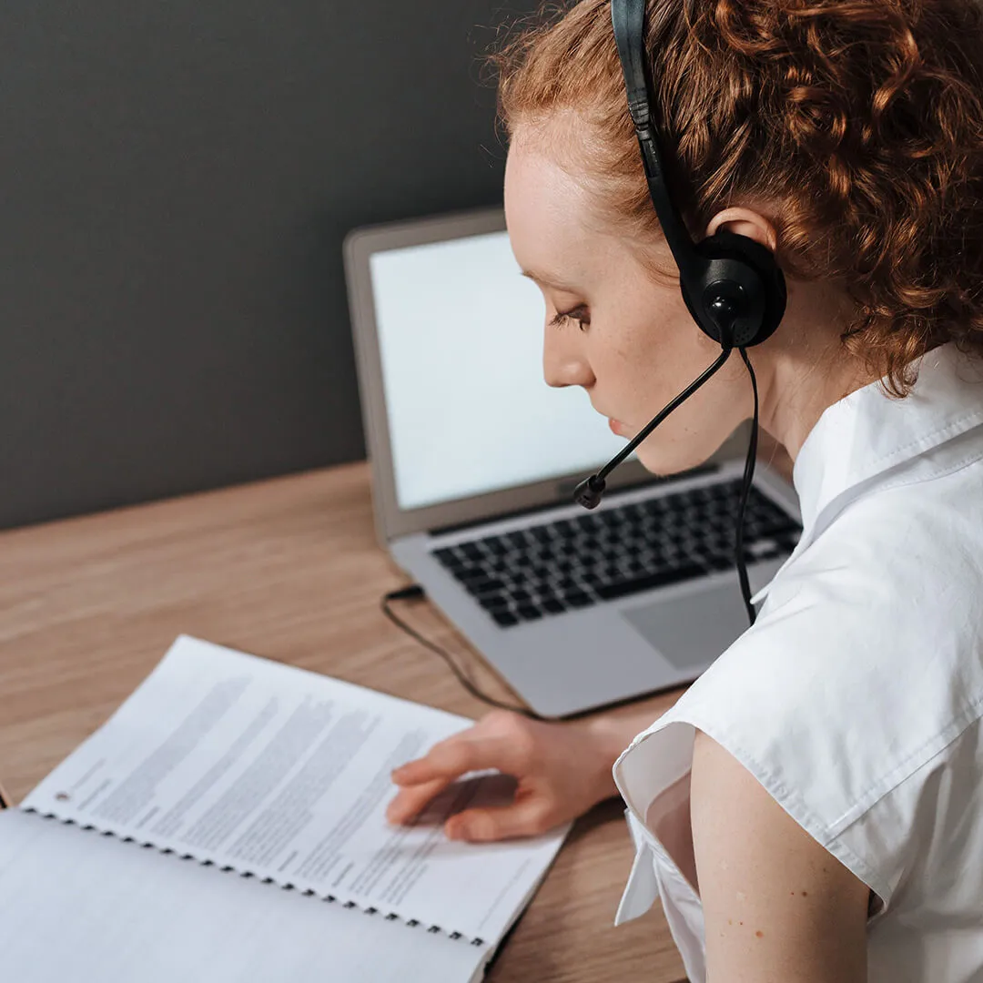 Woman wearing a headset with a microphone while reading a printed document at a desk with a laptop.