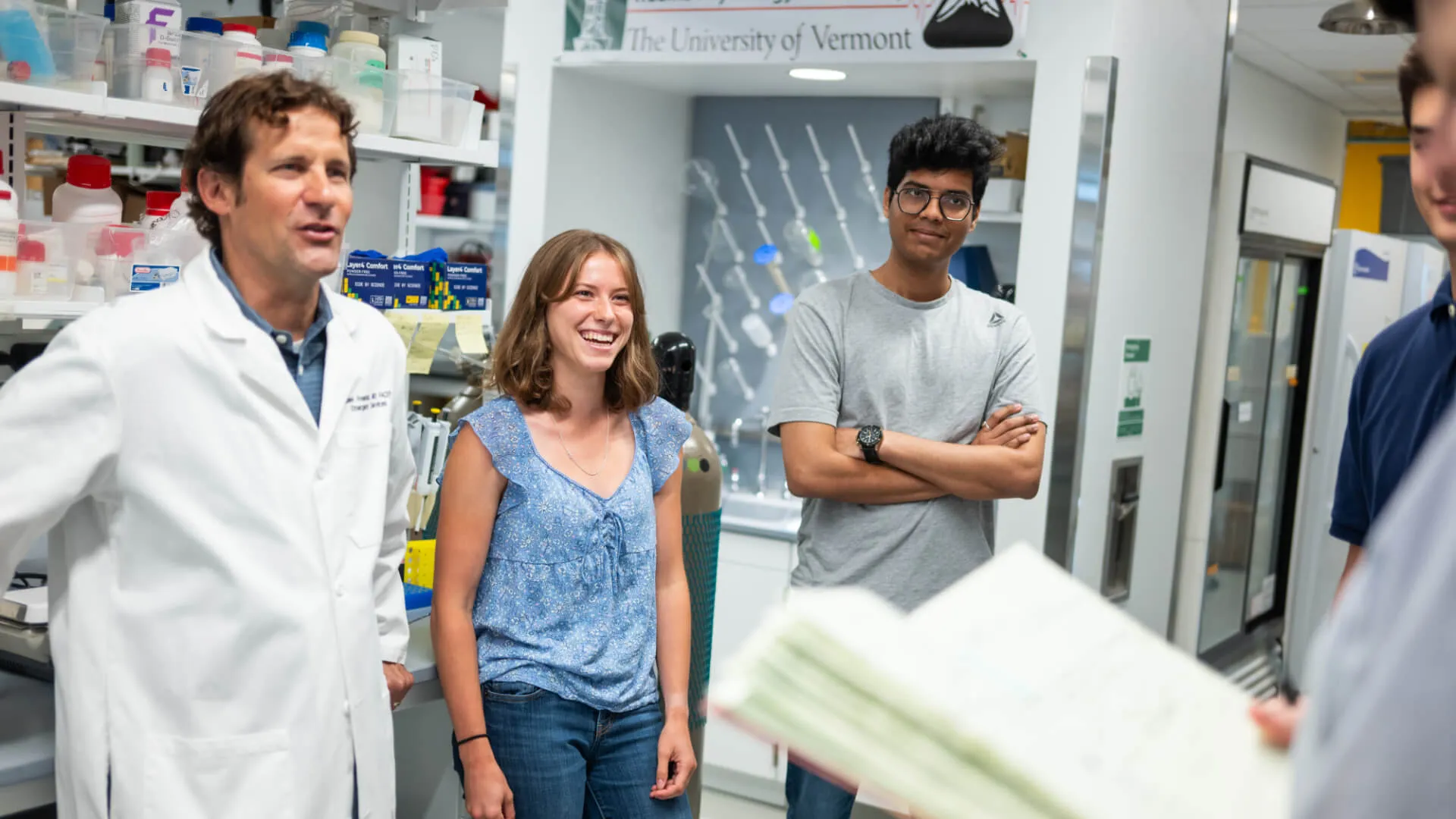 researcher in a lab coat chatting with several students in a lab