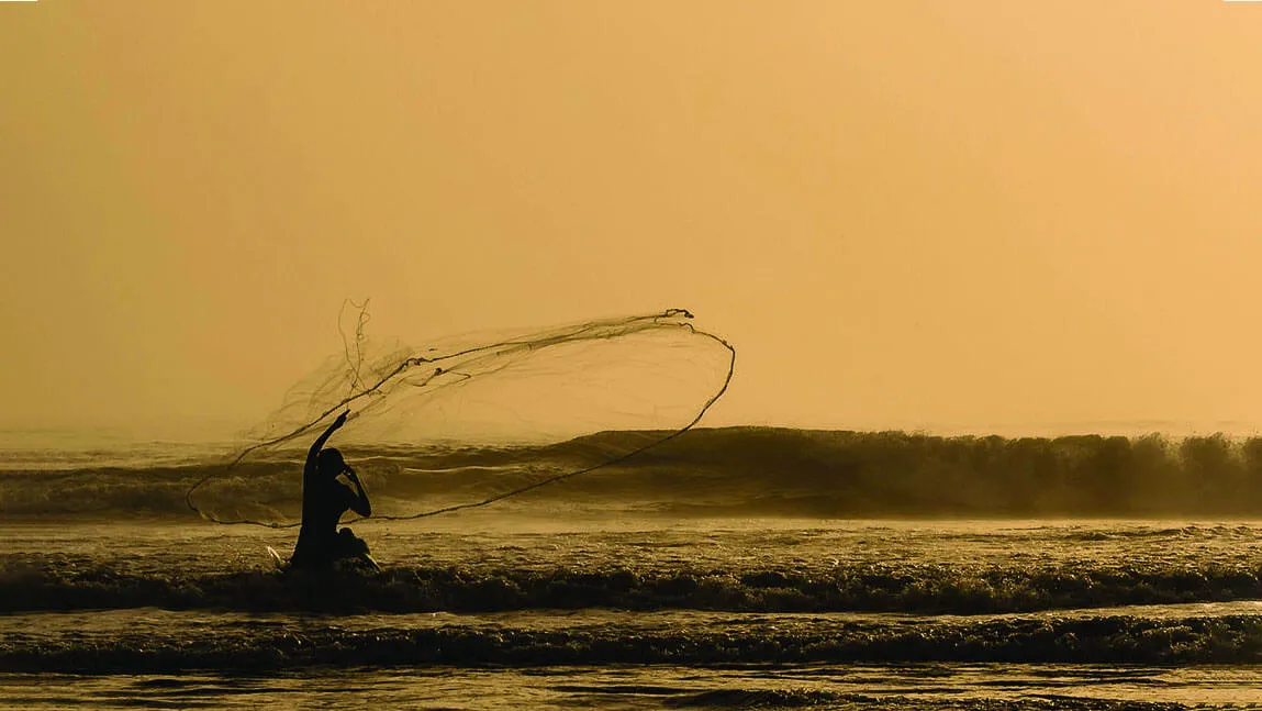 A person silhouetted against an orange sunset casts a wide net into the ocean