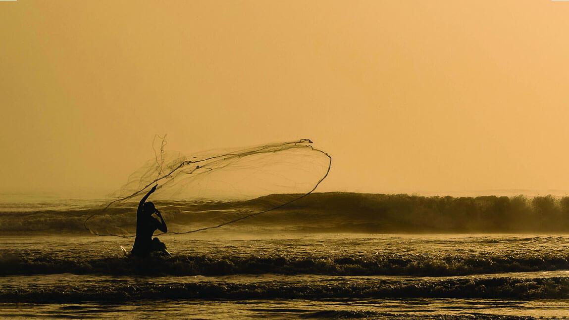 A person silhouetted against an orange sunset casts a wide net into the ocean