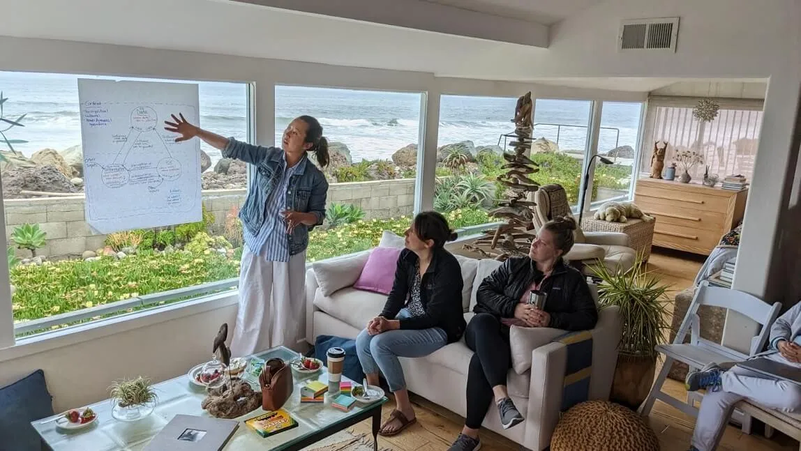 Image of a brainstorming session: Two people sitting on a couch and one person standing pointing to notes on a large poster board. Ocean view in the background.