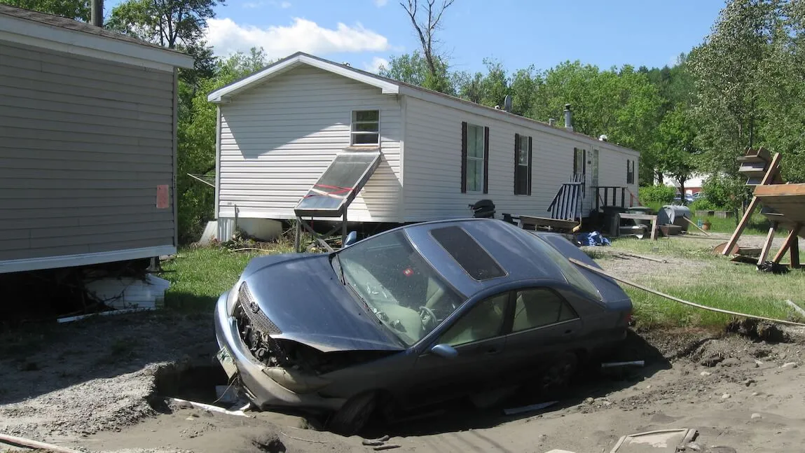 damaged car washed up in the yard of a mobile home resident with more storm debris in the background.