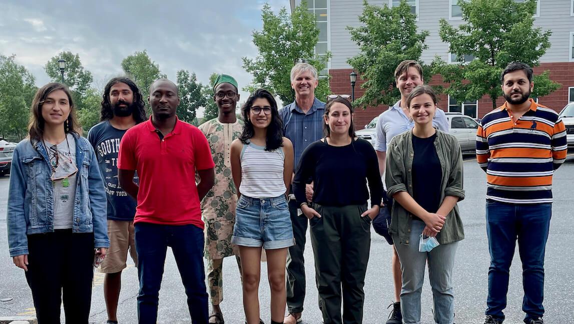 Ten adults standing in a posed photo with trees and buildings in the background.