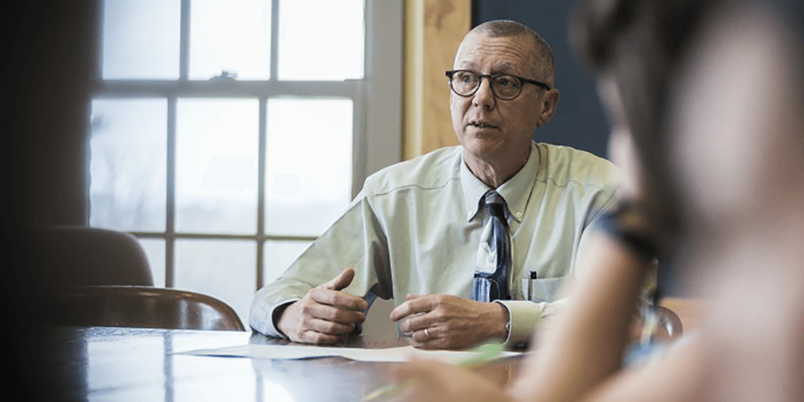 Bob Taylor seated at a table talking with others with light streaming in a window behind him