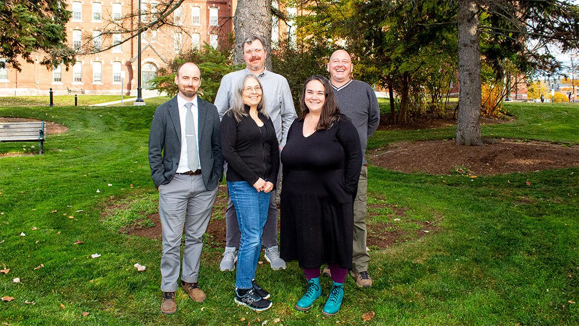 3 male and 2 female professors standing together under a tree on the UVM Green in fall