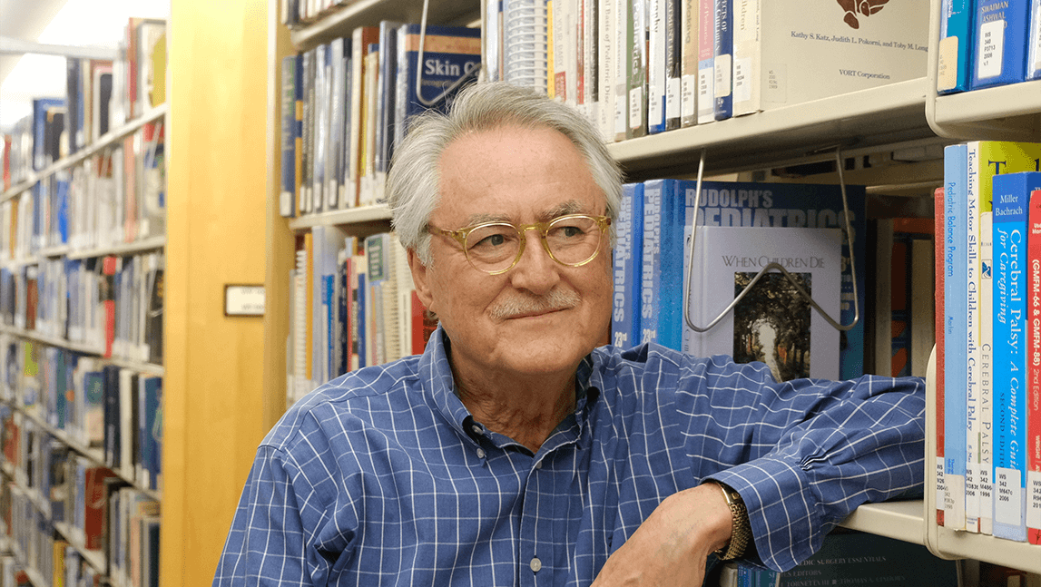 Allan Kellehear leaning on a shelf of books in a library like setting