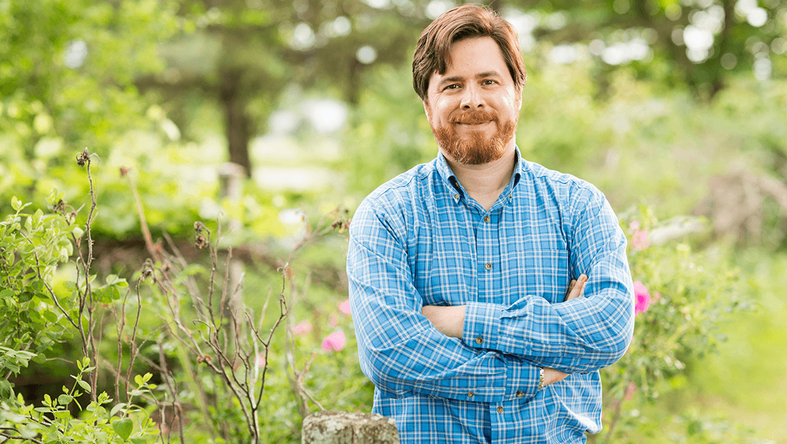 Peter Henne, standing in a field with grass and flowers with a wooden fence in the background