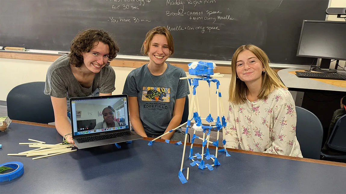 three students (plus one on video) sitting at table in classroom with project in front of them, smiling toward camera