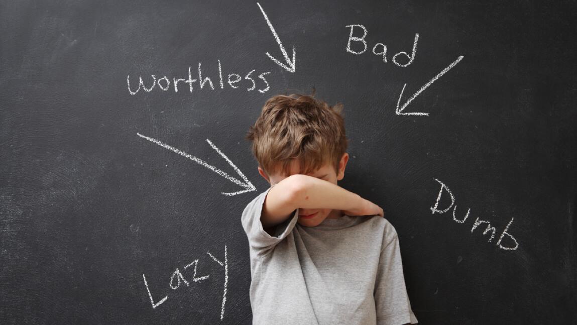 Child covering his eyes and frowning, text that looks like it was written with chalk reads worthless, bad, dumb, lazy, with arrows pointing to child