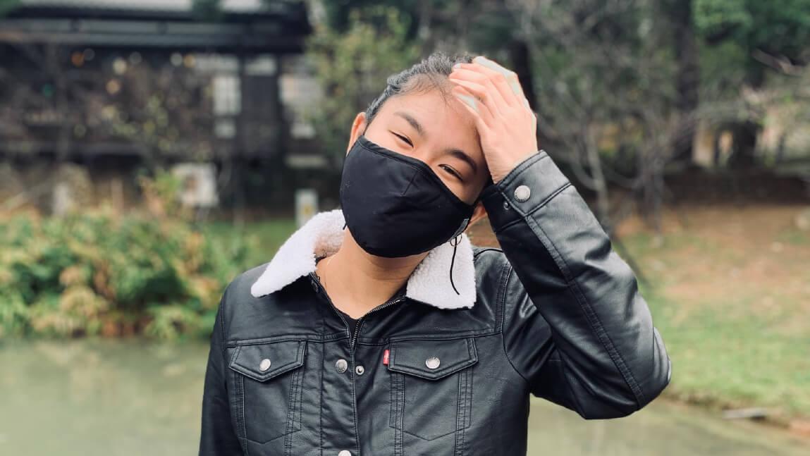 Person standing outside wearing a mask, looking at the camera with her hand on her head and head tilted