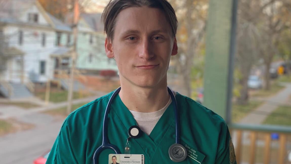 Person outdoors wearing green hospital scrubs, hospital name badge and a stethoscope around their neck