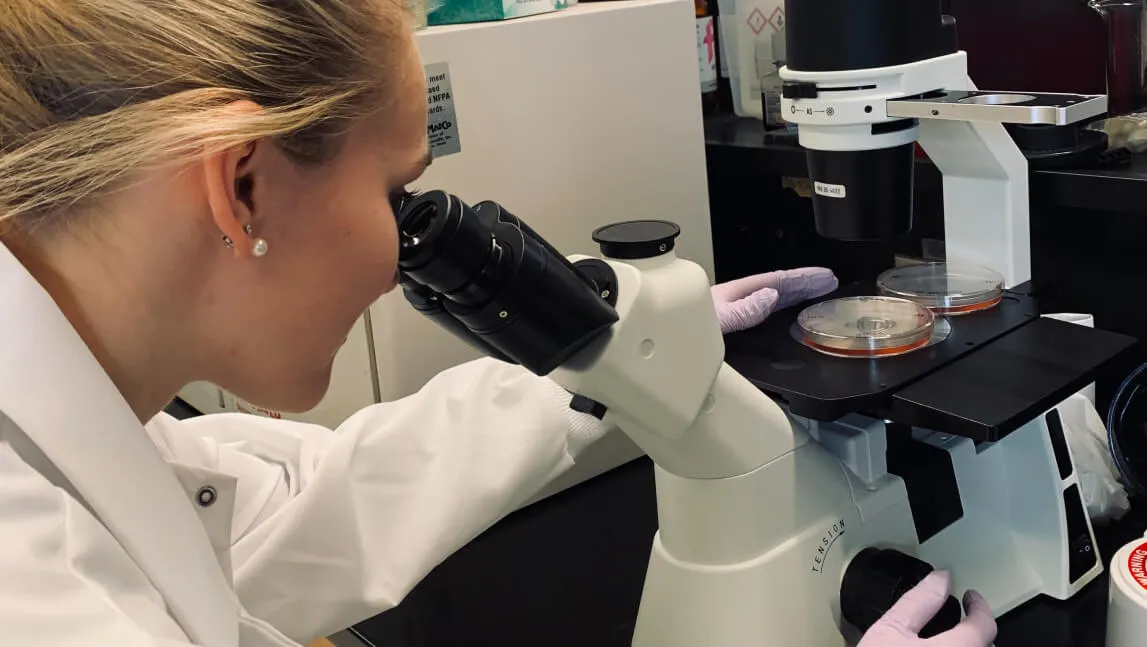 Person in a white lab coat peering through a microscope
