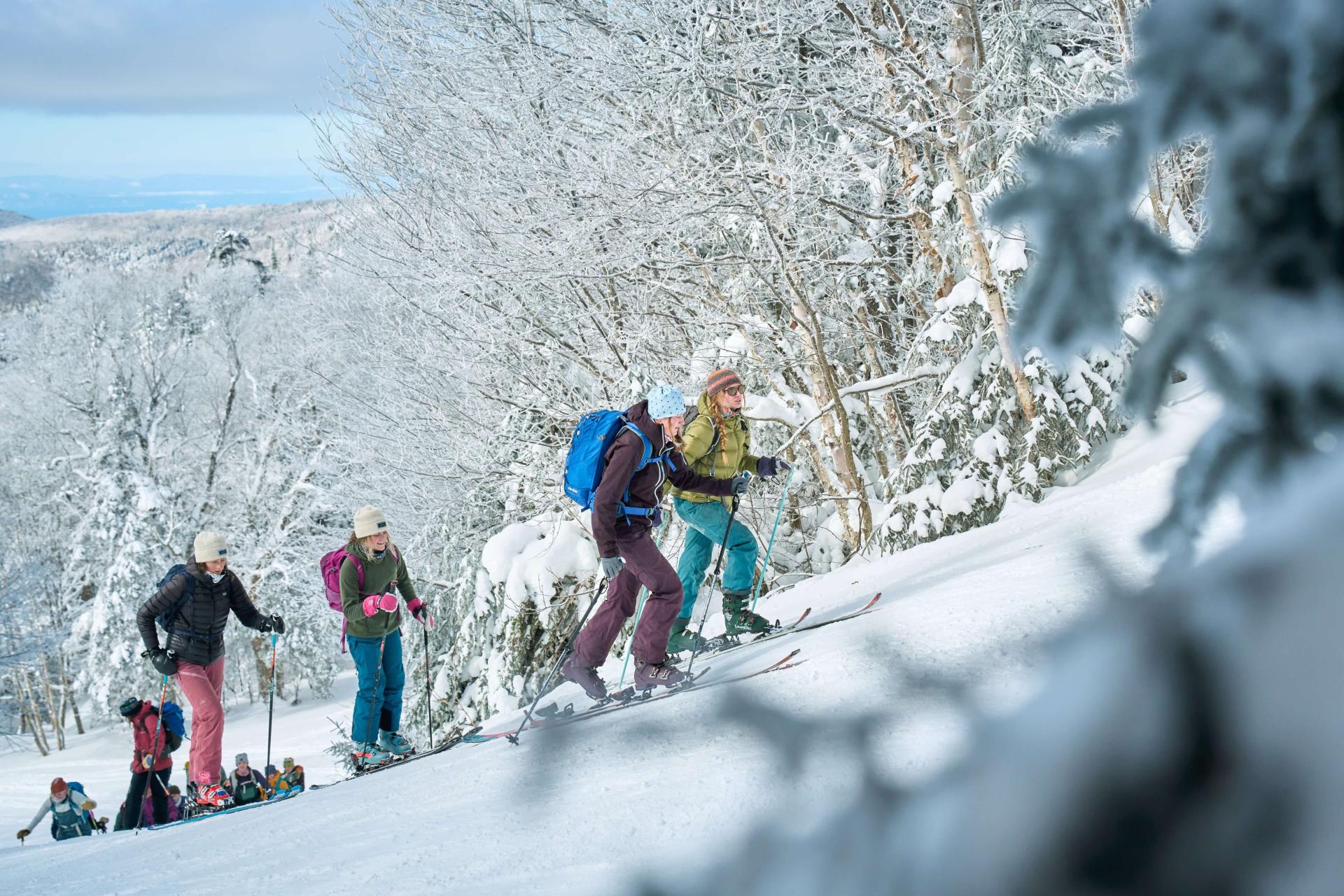 A photo of several people clad in colorful snow gear trekking up a brightly lit, snowy mountain trail. They look enthusiastic and energetic. 