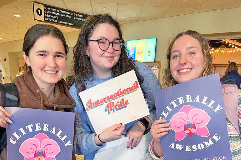 Three students pose for the camera, all holding posters from CHWB's 2023 Sexpo. One poster says "Cliterally Awesome" and the other says "Intersectional Pride"