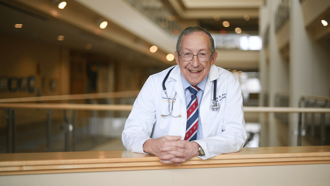 a smiling man in a white coat leaning on a balcomy wall