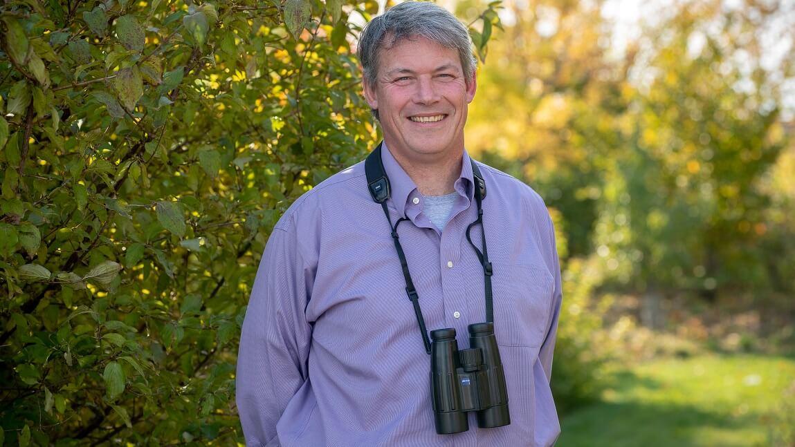 Allan Strong outside in front of greenery with binoculars around his neck.
