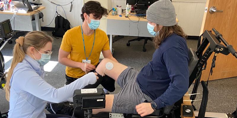 Two people wearing masks place electrical pads on the legs of a third person wearing a mask and seated in a rowing machine.