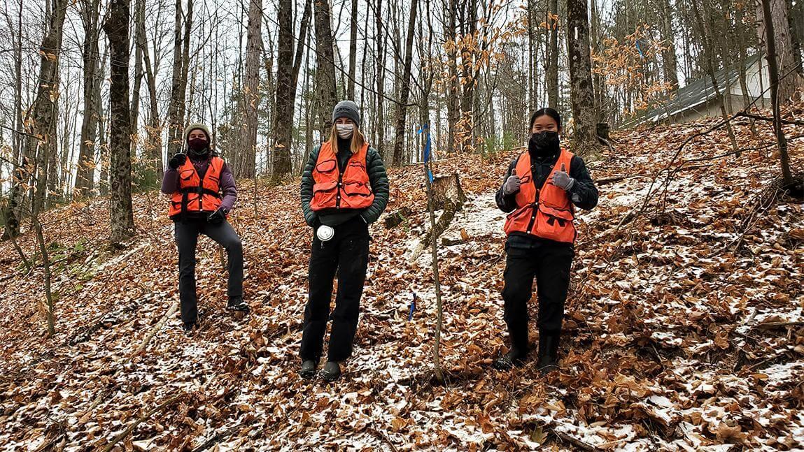 Three students in orange vests and face masks stand in forest in the fall