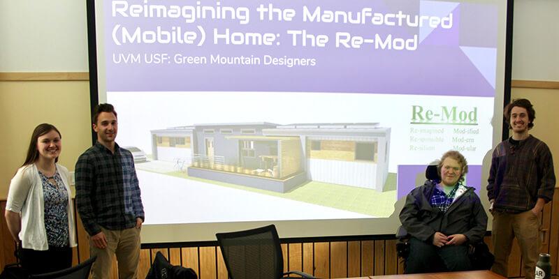 Students stand next to screen showing presentation of their design.