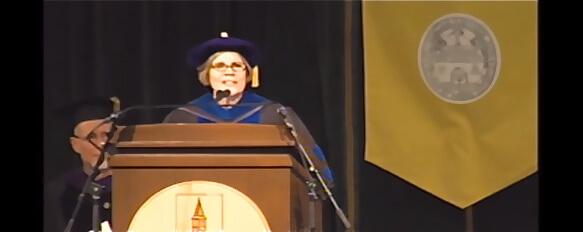 Speaker in regalia at podium. Yellow banner on the right.