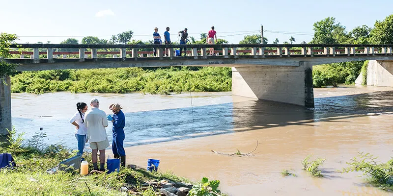 Researchers stand on bank of river in Cuba with onlookers on bridge