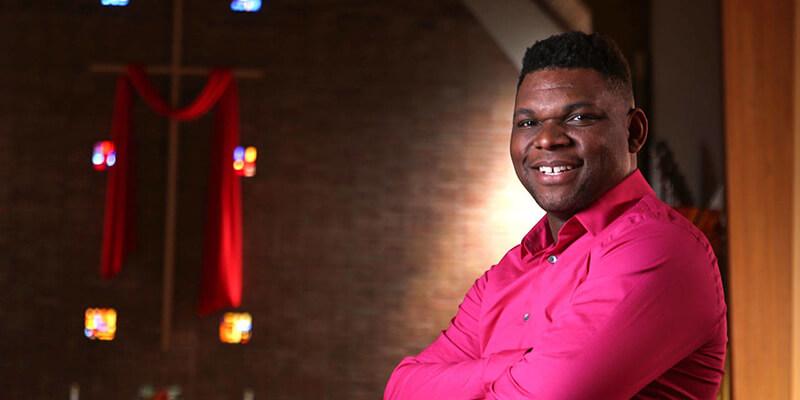 Rev. Darrell Goodwin stands in church, with arms crossed in pink shirt, smiling to camera